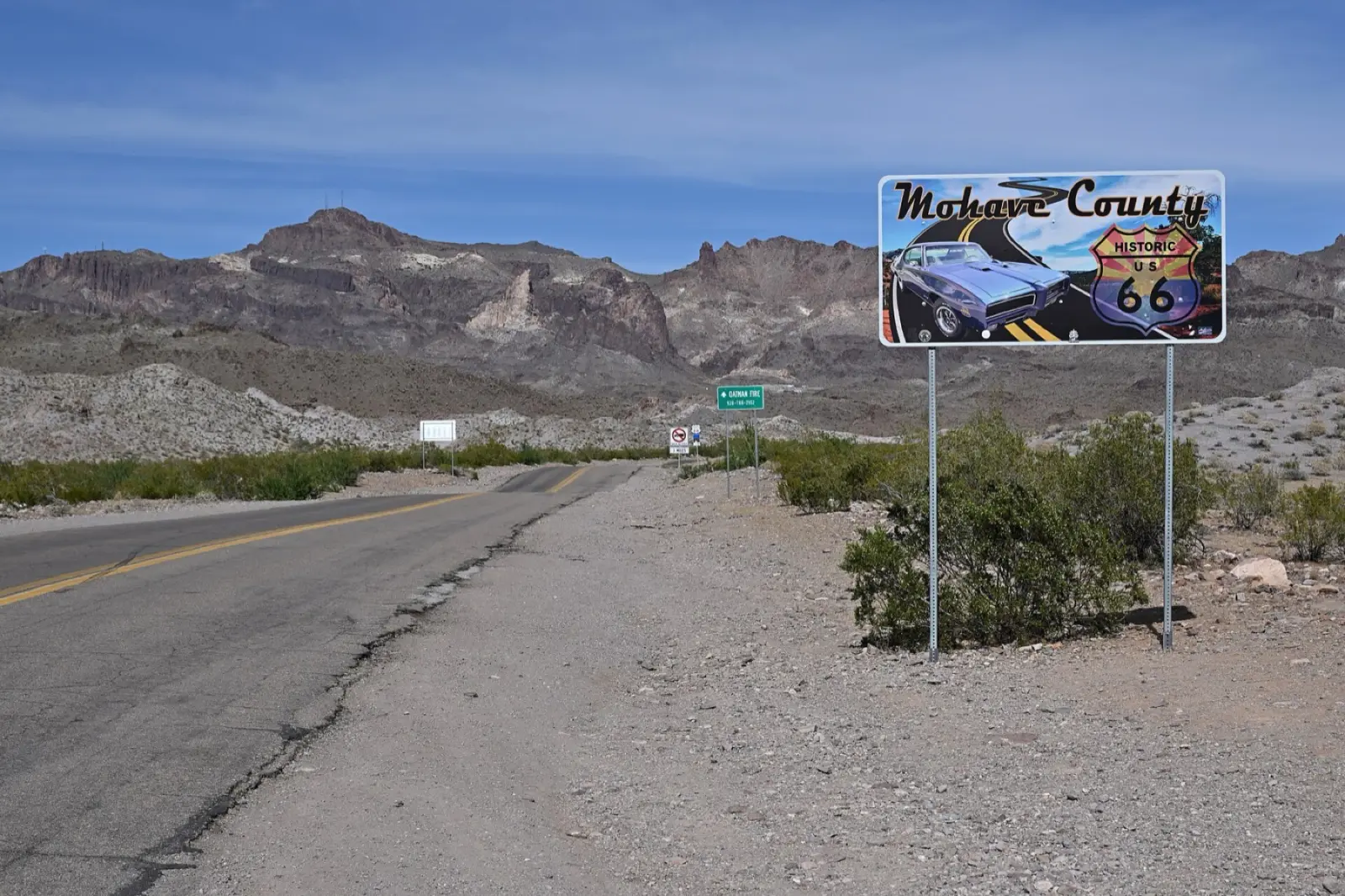 A Mohave County Historic US 66 sign beside the two-lane Oatman Highway, with desert scrub and jagged mountains behind