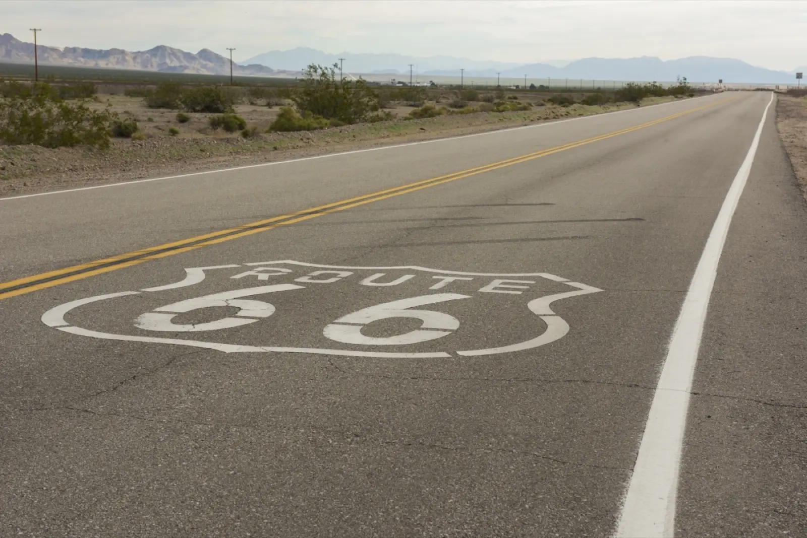 Route 66 shield painted on the asphalt of a straight two-lane desert road, with creosote bushes and bare mountains in the distance