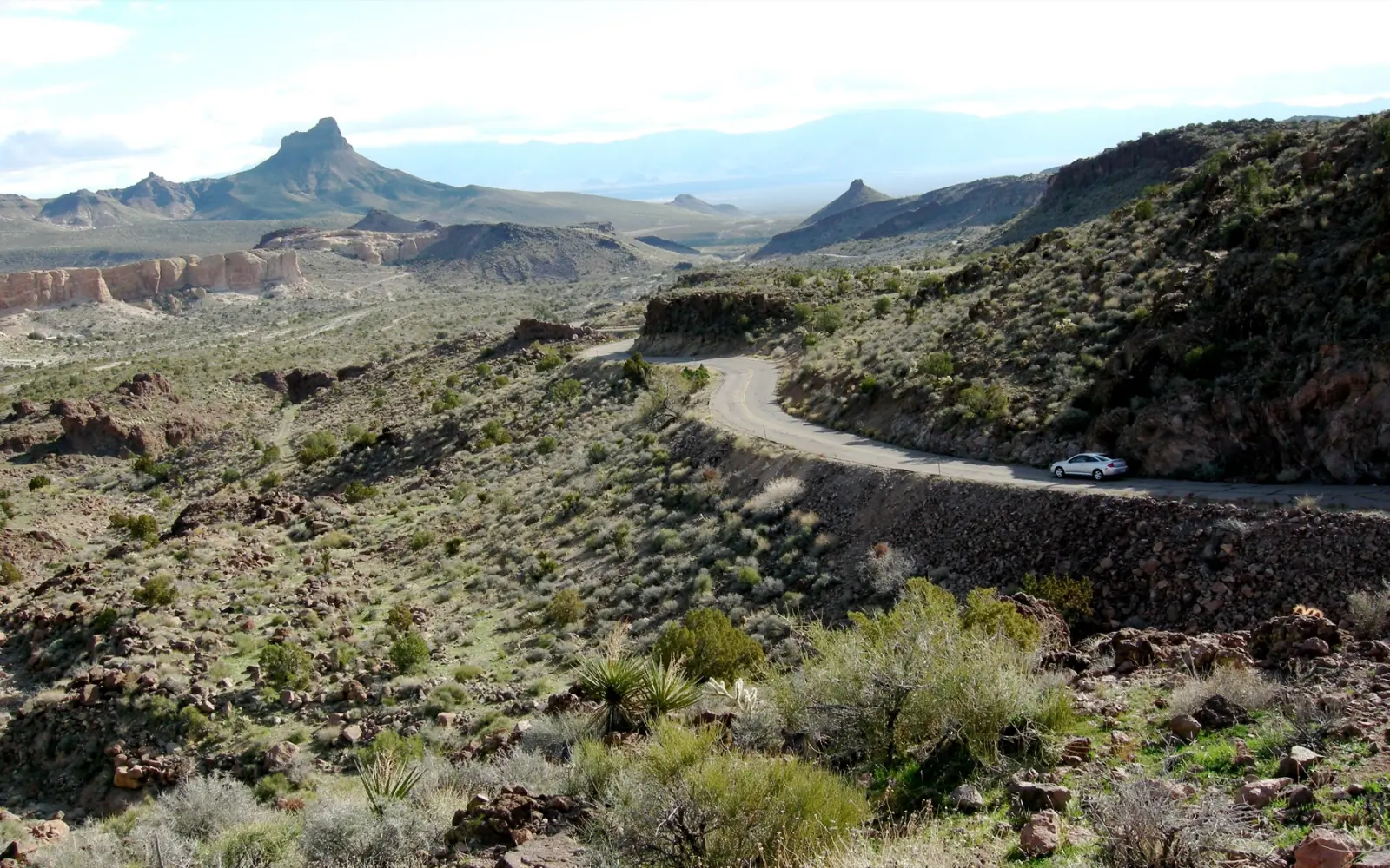 A silver car on a narrow paved curve of old Route 66 cut into a rocky hillside in the Black Mountains of Arizona, with no guardrail and desert peaks beyond