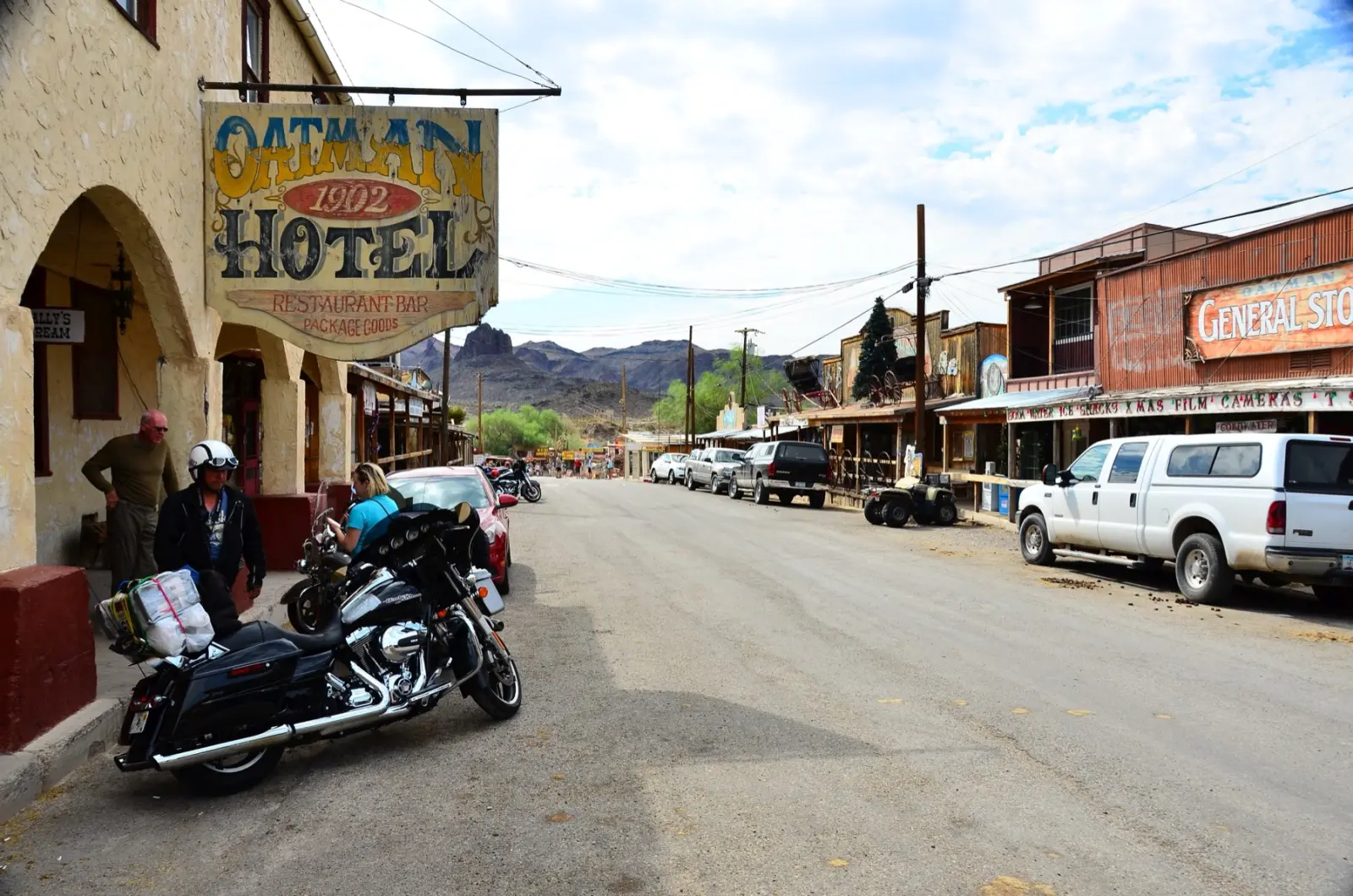 Main street of Oatman, Arizona, with the 1902 Oatman Hotel sign, a parked touring motorcycle, pickup trucks and the Black Mountains behind
