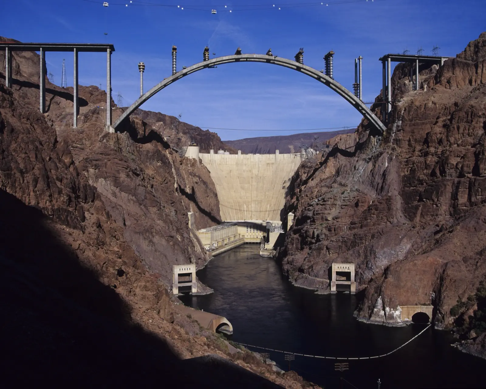 The arch of the Hoover Dam bypass bridge under construction high above the Colorado River, with Hoover Dam behind it