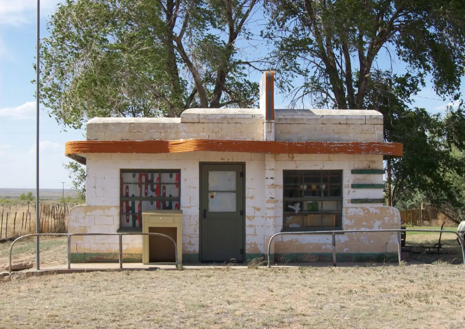 Small abandoned roadside cafe in Glenrio with peeling white paint, an orange curved awning, a green door and big cottonwood trees behind it