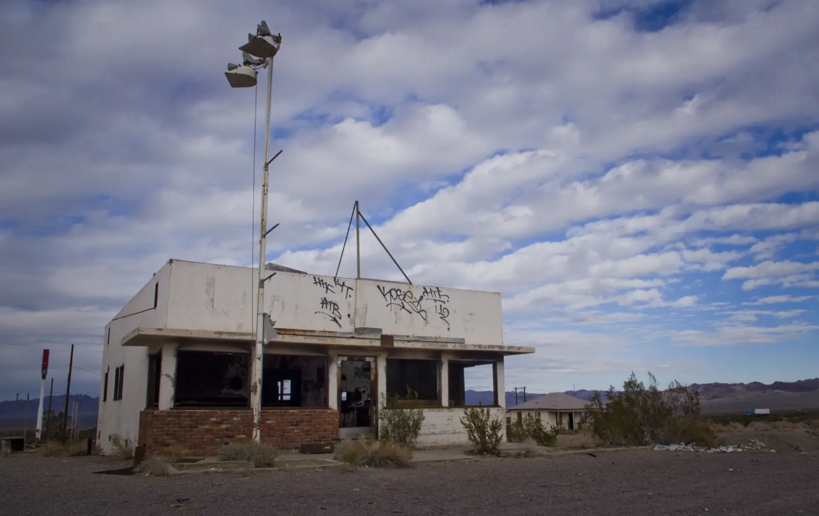 Abandoned white cafe building with graffiti, broken windows and a tall light pole on the old road in Ludlow, California, under a cloudy desert sky