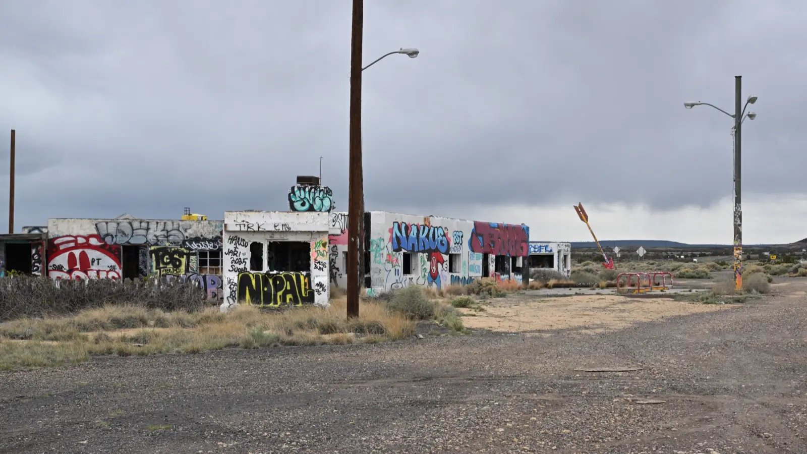 Abandoned Twin Arrows Trading Post buildings covered in graffiti beside old Route 66 in Arizona, with one tall red and yellow arrow standing at an angle under a gray sky