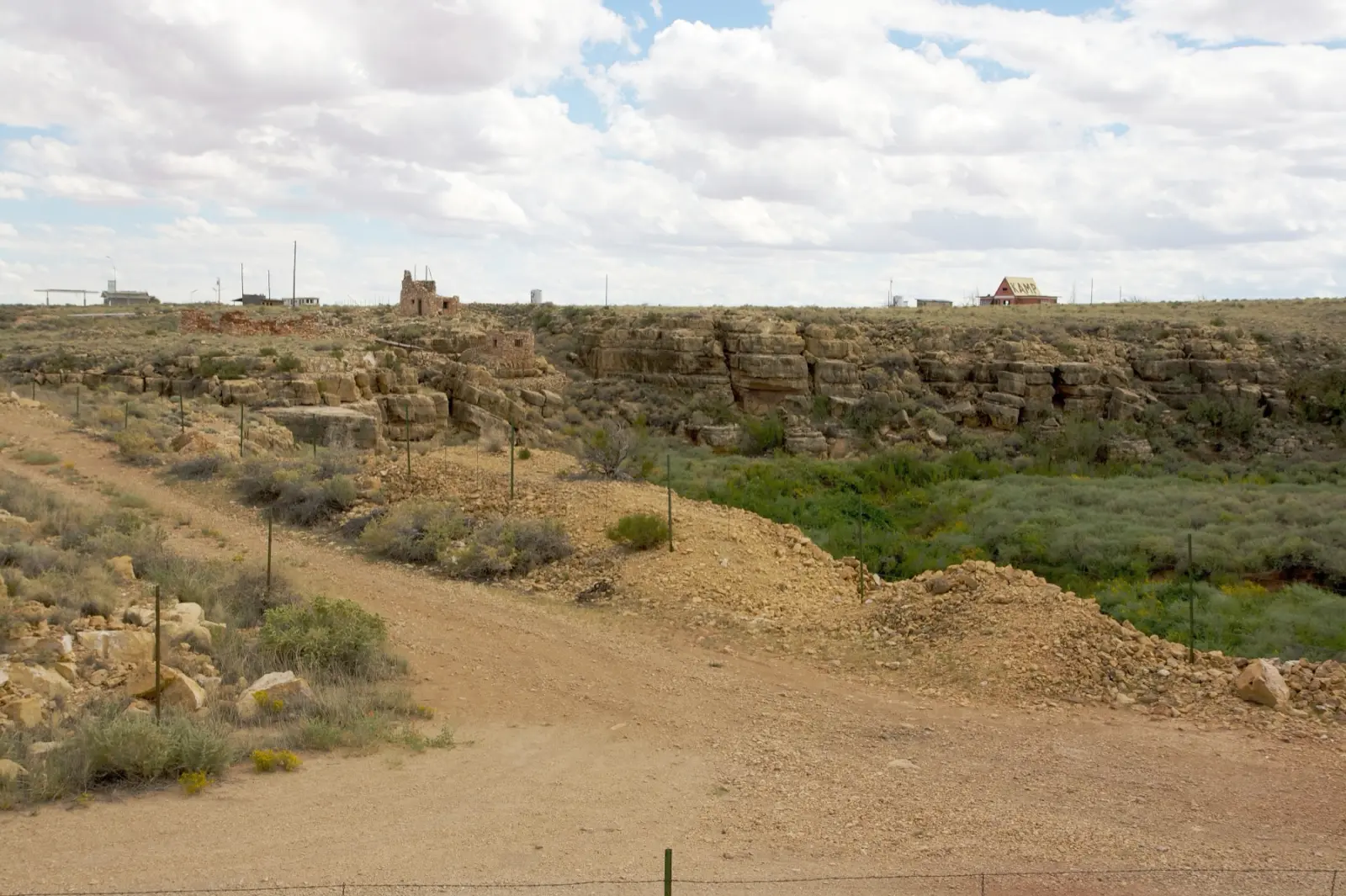Stone ruins of Two Guns on the rim of Canyon Diablo in Arizona, with a dirt road in front and an abandoned red building with the word CAMP on the roof in the distance