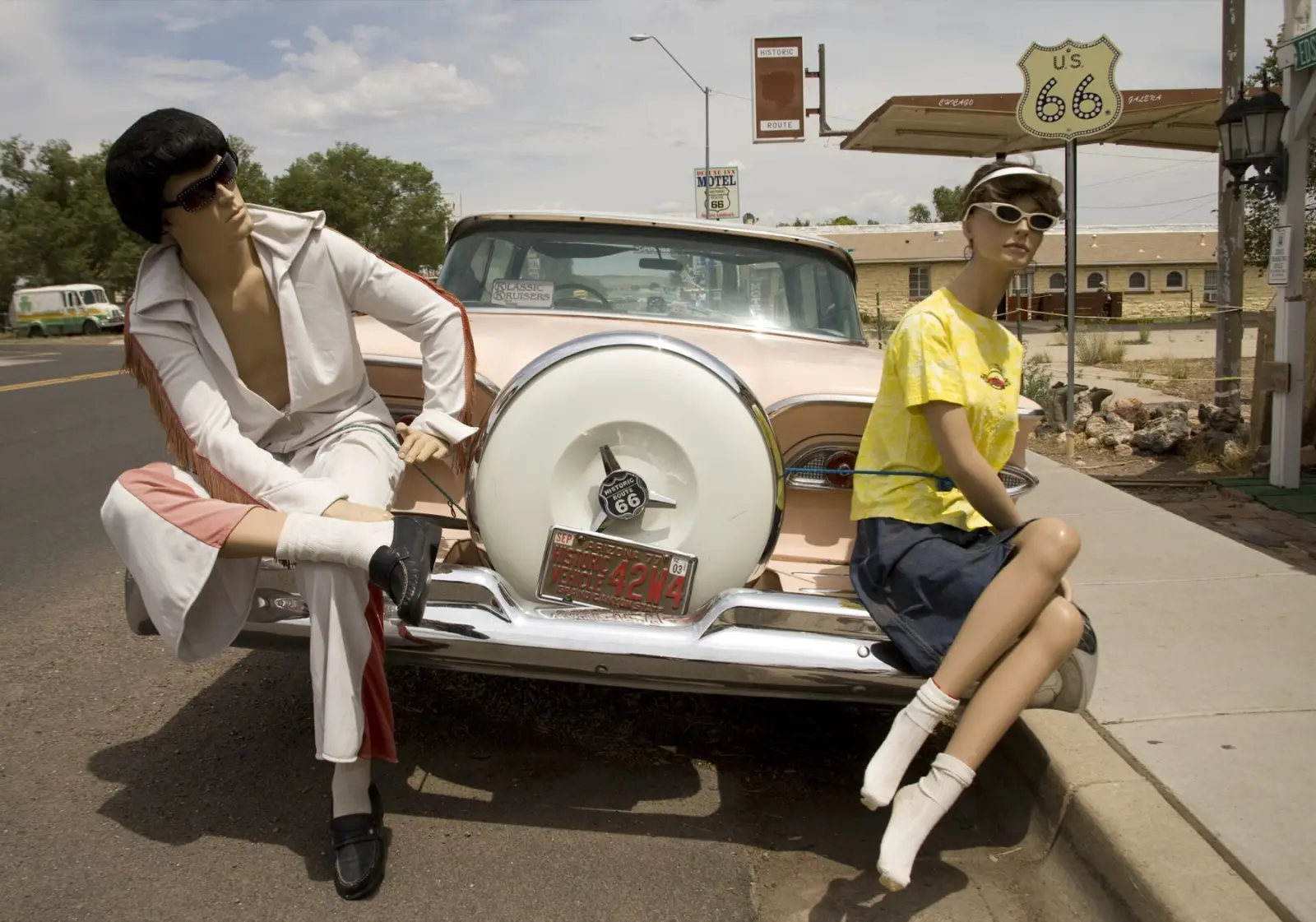 Mannequins dressed in 1950s style sitting on the bumper of a pink vintage car under a US 66 shield sign in Seligman, Arizona