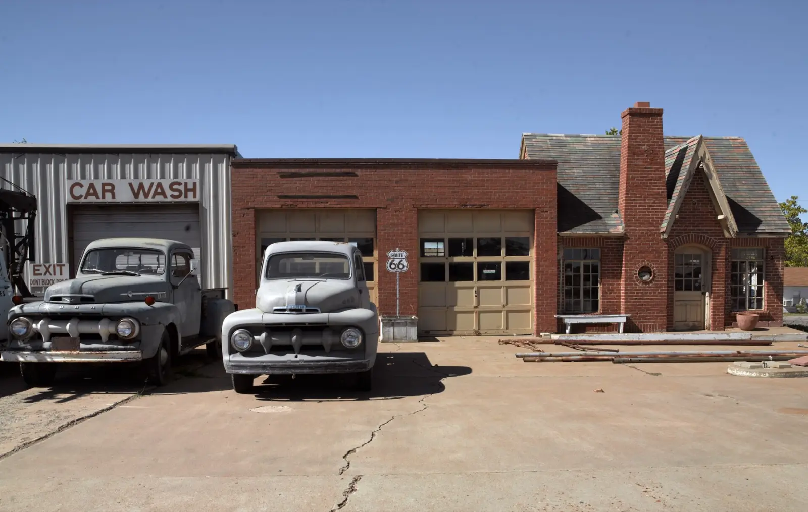 Restored red brick Phillips 66 station with a cottage-style roof and chimney in Chandler, Oklahoma, with two old gray Ford pickup trucks parked in front and a Route 66 shield sign