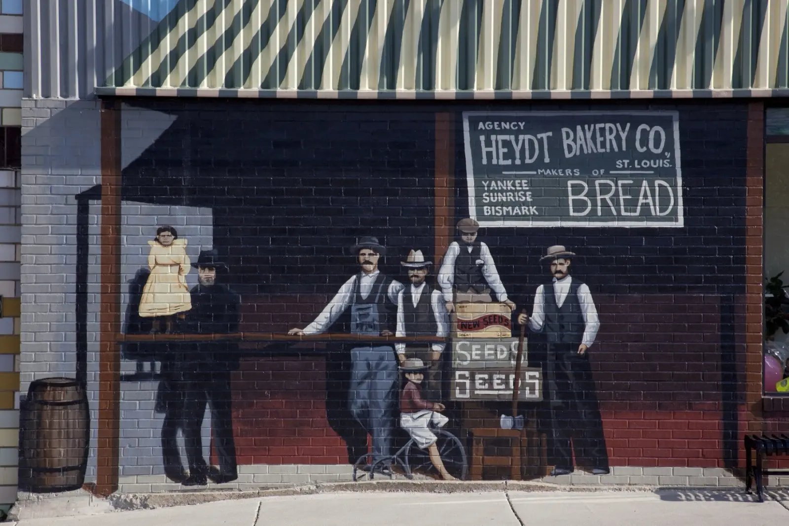 Painted mural on a brick wall in Cuba, Missouri, showing men in vests and hats outside a store with a Heydt Bakery Co. bread sign and a crate of seeds