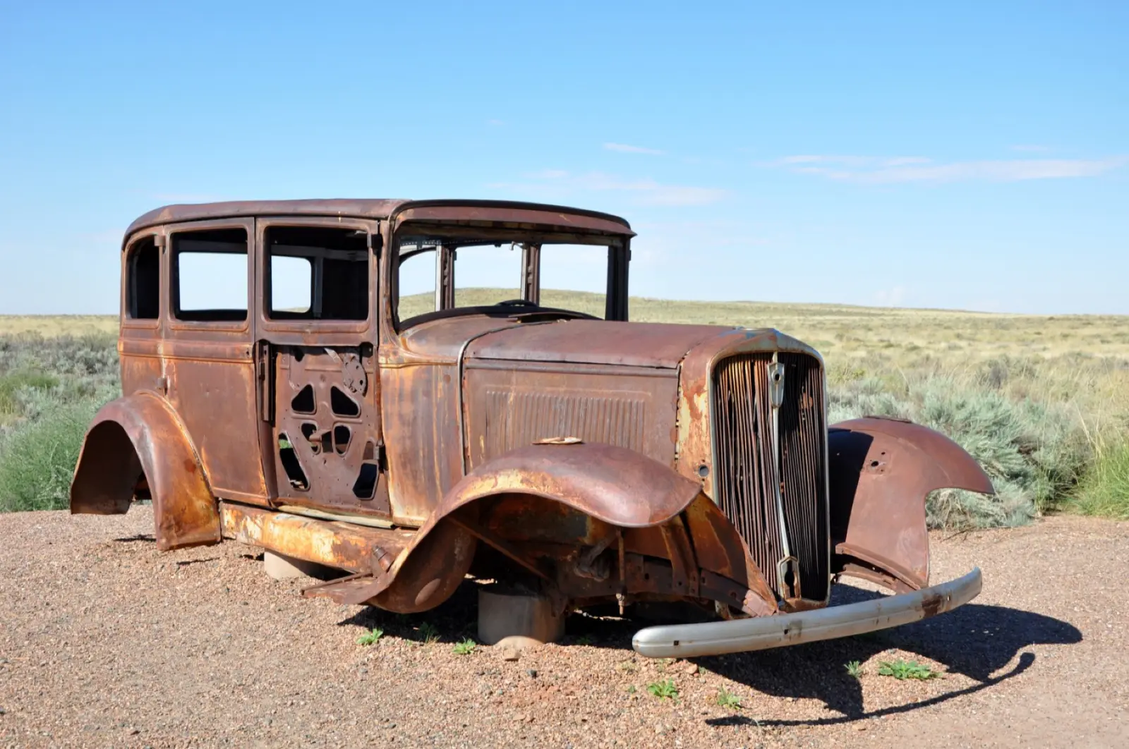 Rusted 1930s Studebaker sedan on gravel beside dry grassland under a blue sky