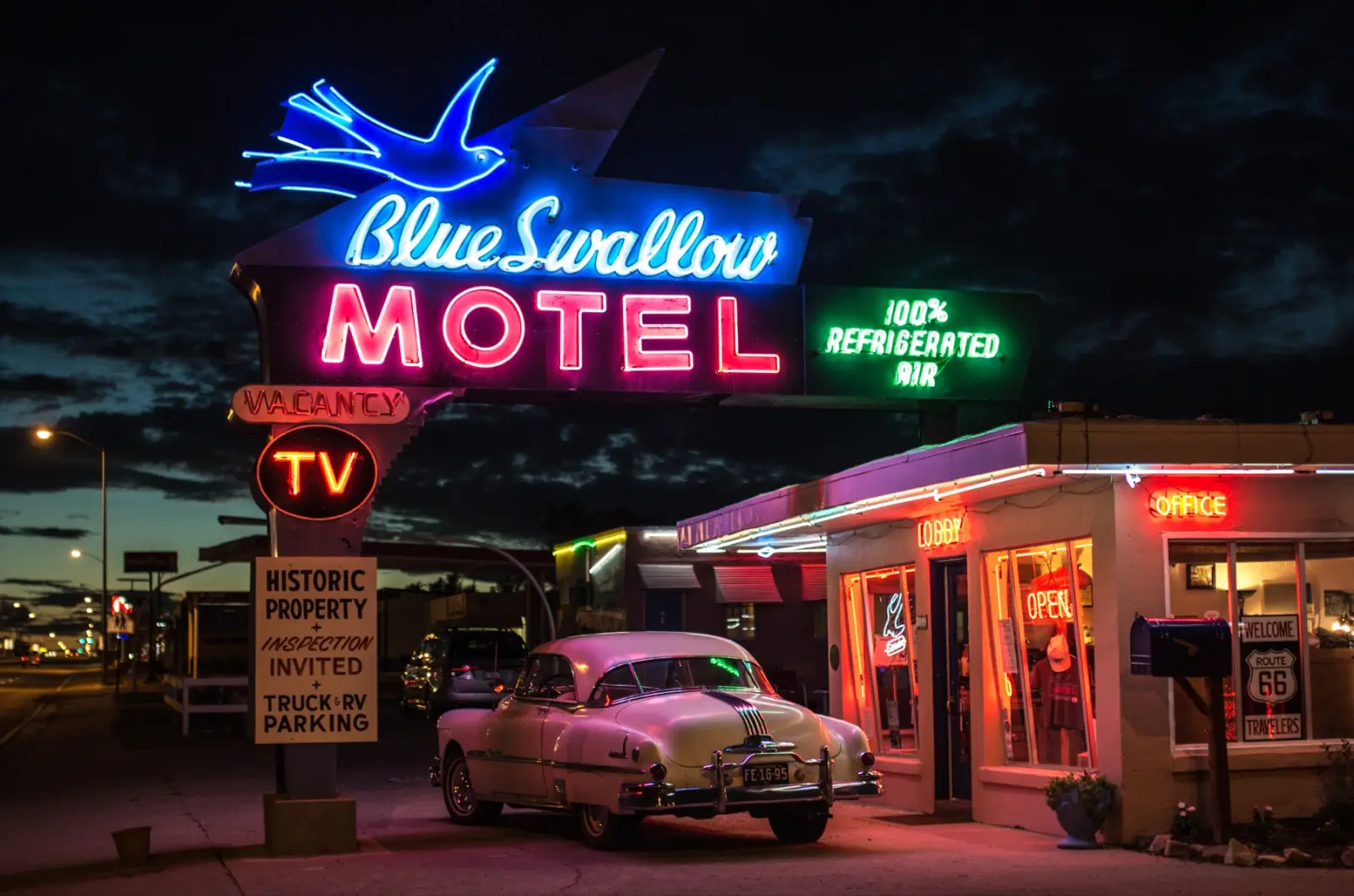 Blue Swallow Motel neon sign glowing at dusk in Tucumcari, New Mexico, with a cream 1950s car parked by the lit office