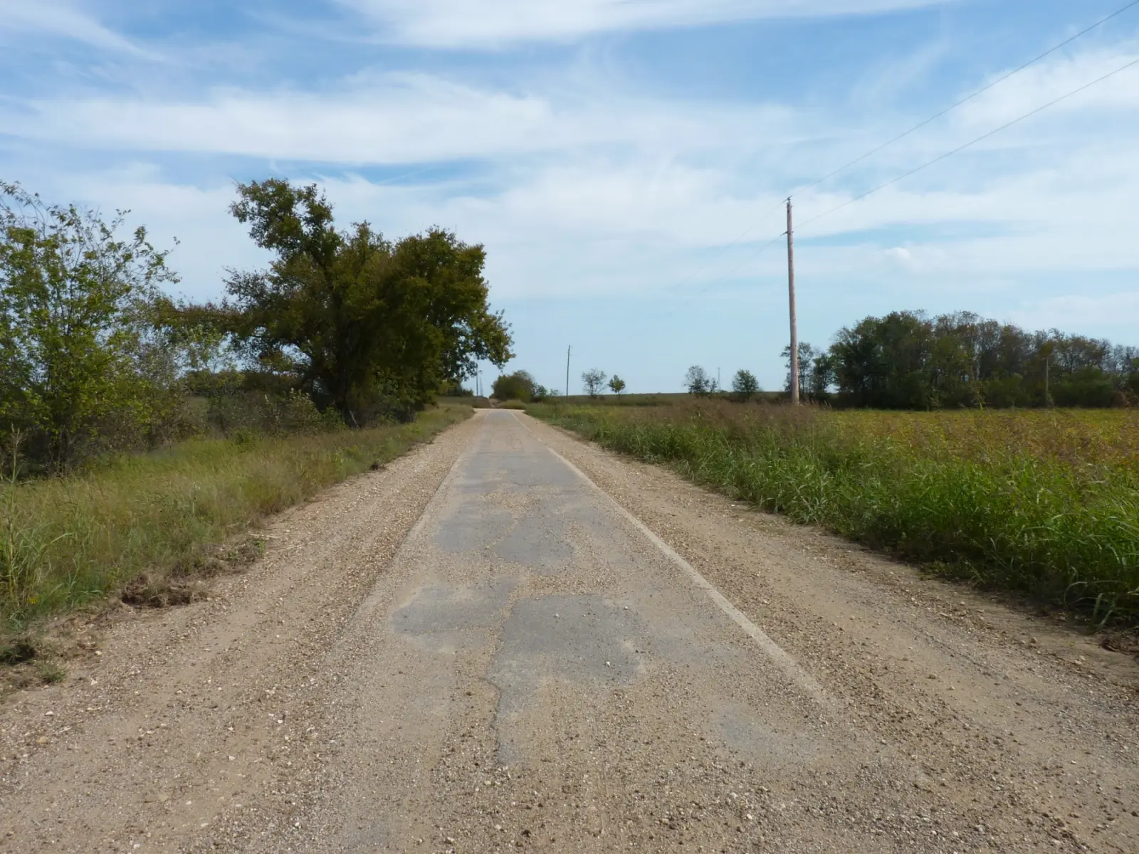 Narrow strip of old cracked pavement with gravel shoulders running straight between fields and trees near Miami, Oklahoma, under a blue sky