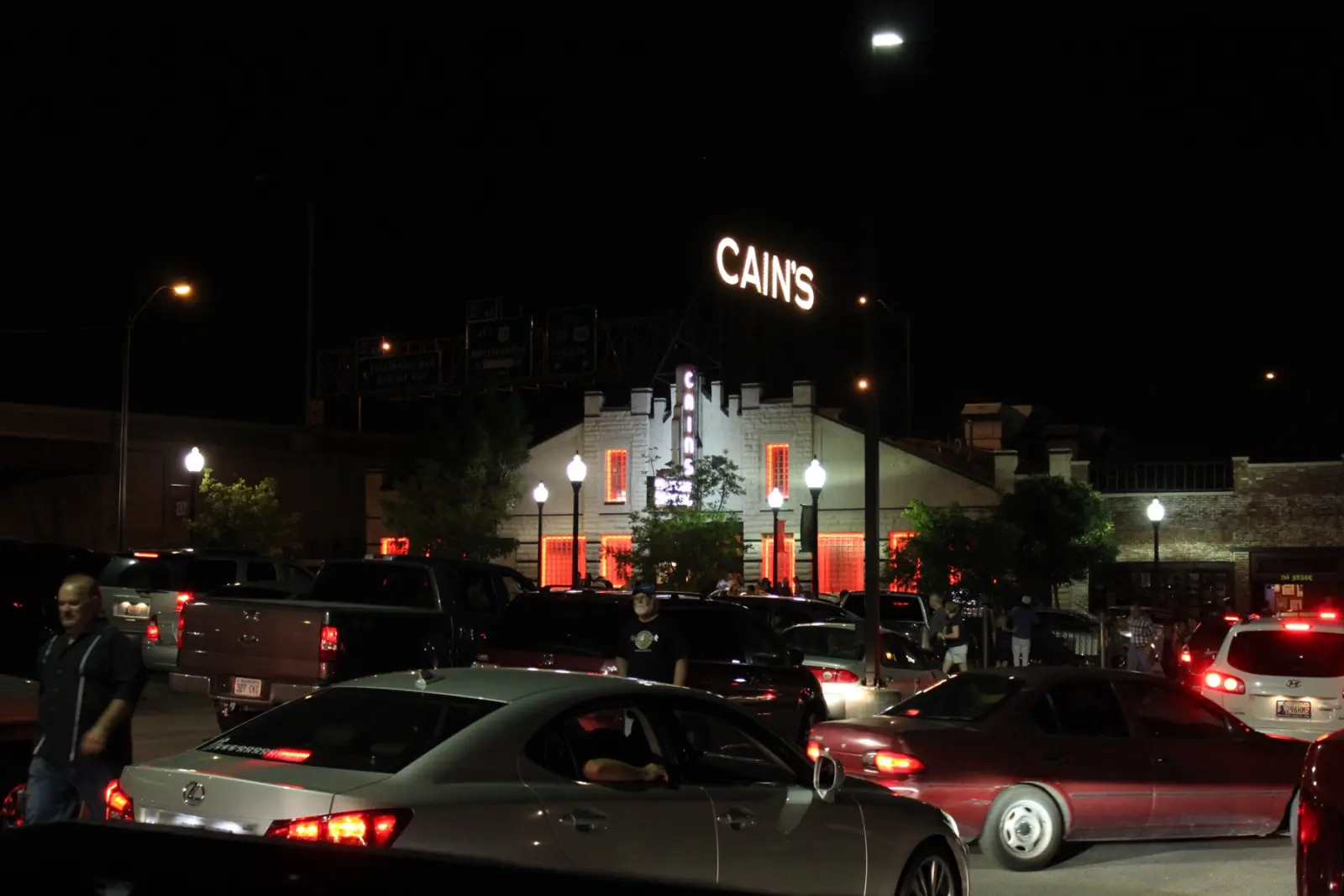 Cain's Ballroom in Tulsa at night, its neon CAIN'S sign lit above the white brick building, with a crowd and cars in the lot under the highway overpass signs