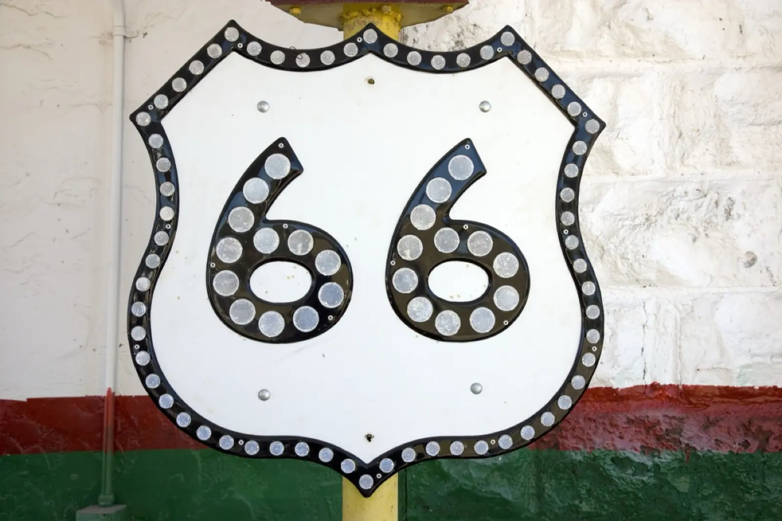 A vintage Route 66 shield sign studded with reflectors, mounted on a yellow post against a white wall