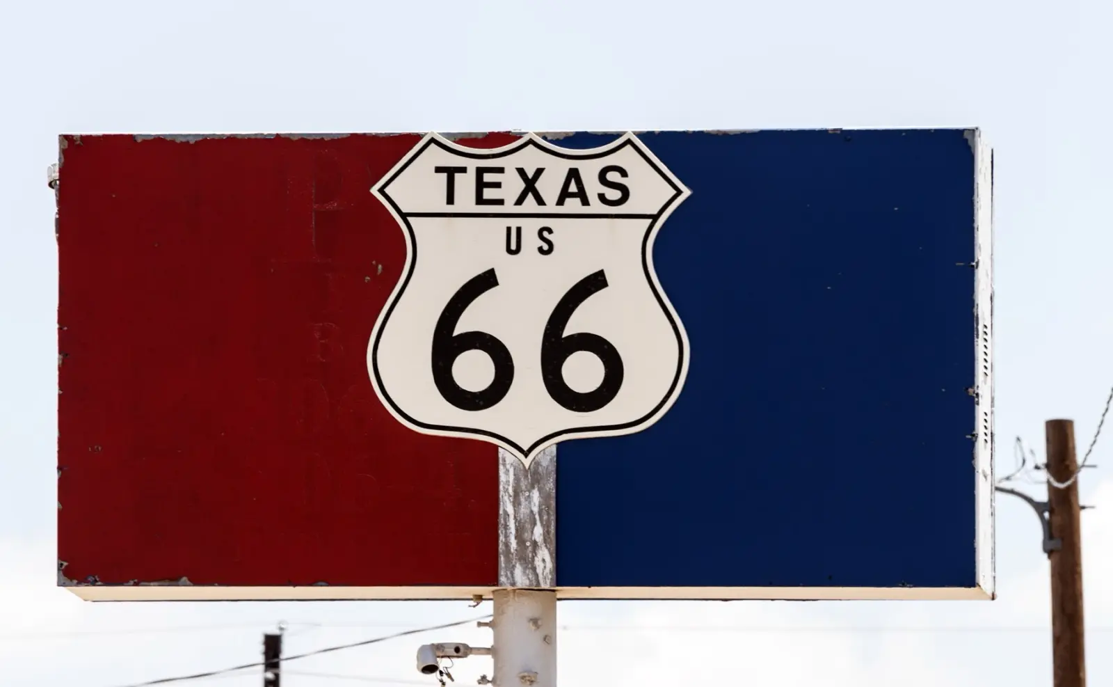 A white Texas US 66 highway shield mounted on a faded red and blue sign on a pole against a pale sky in Amarillo, Texas